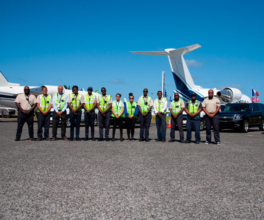 This image brings to life a scene at ExecuJet's St. Maarten Fixed-Base Operator (FBO), focusing on the ground handling team ready for action on the runway. Envision a group of 13 individuals, both men and women, standing together, each adorned in reflective gear that glimmers and catches the light. The fabric of their uniforms feels crisp and efficient, mirroring their readiness to attend to the aircraft. Behind them, the parked aircraft stand silent and grand, waiting for the team's expert care, and a number of SUVs are strategically positioned nearby, their engines humming in preparation. The whole scene conveys a sense of organization, teamwork, and the vibrant behind-the-scenes world of aviation.